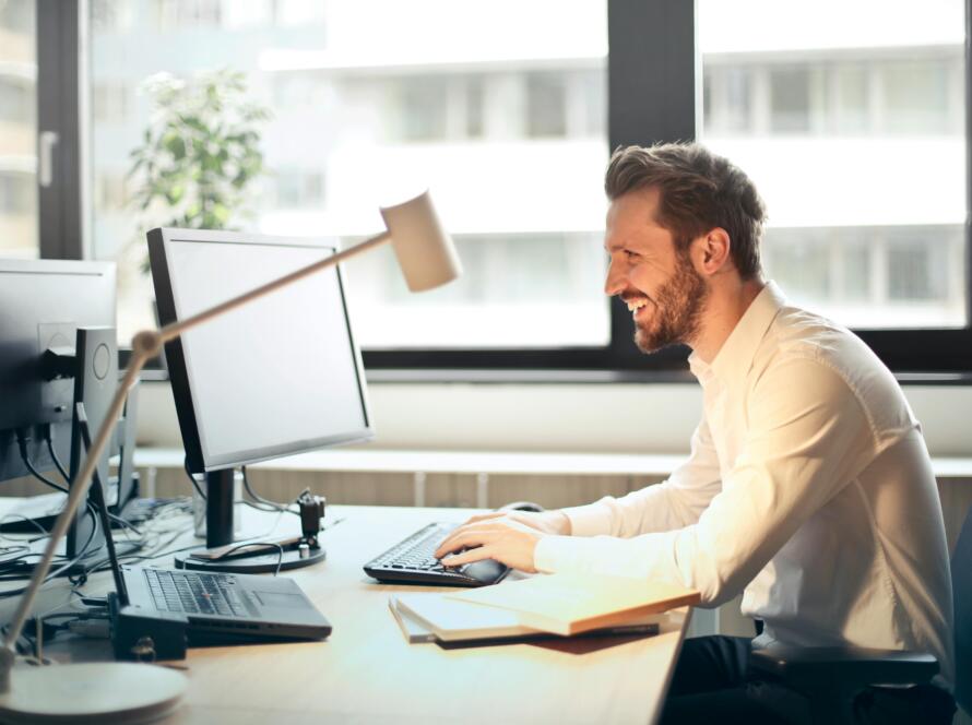 A man smiling while working at an office desk with a computer and natural daylight streaming in through large windows.